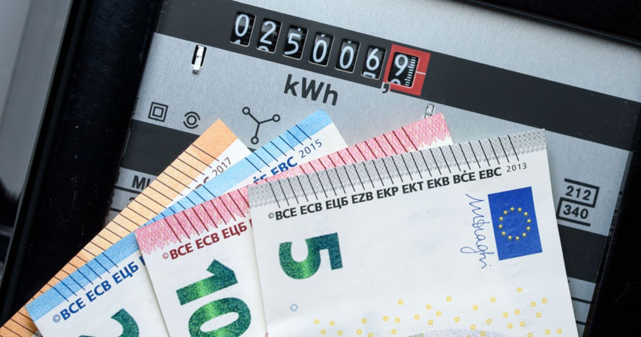 Abgebildet ist ein Stromzähler, davor werden verschiedene Geldscheine gehalten.