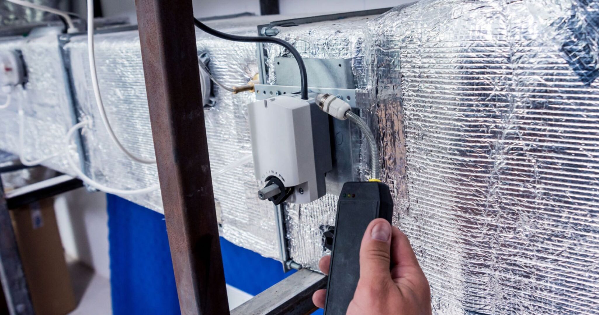 Eine Hand hält einen Gassensor an eine Gasleitung in einem Kellerraum.