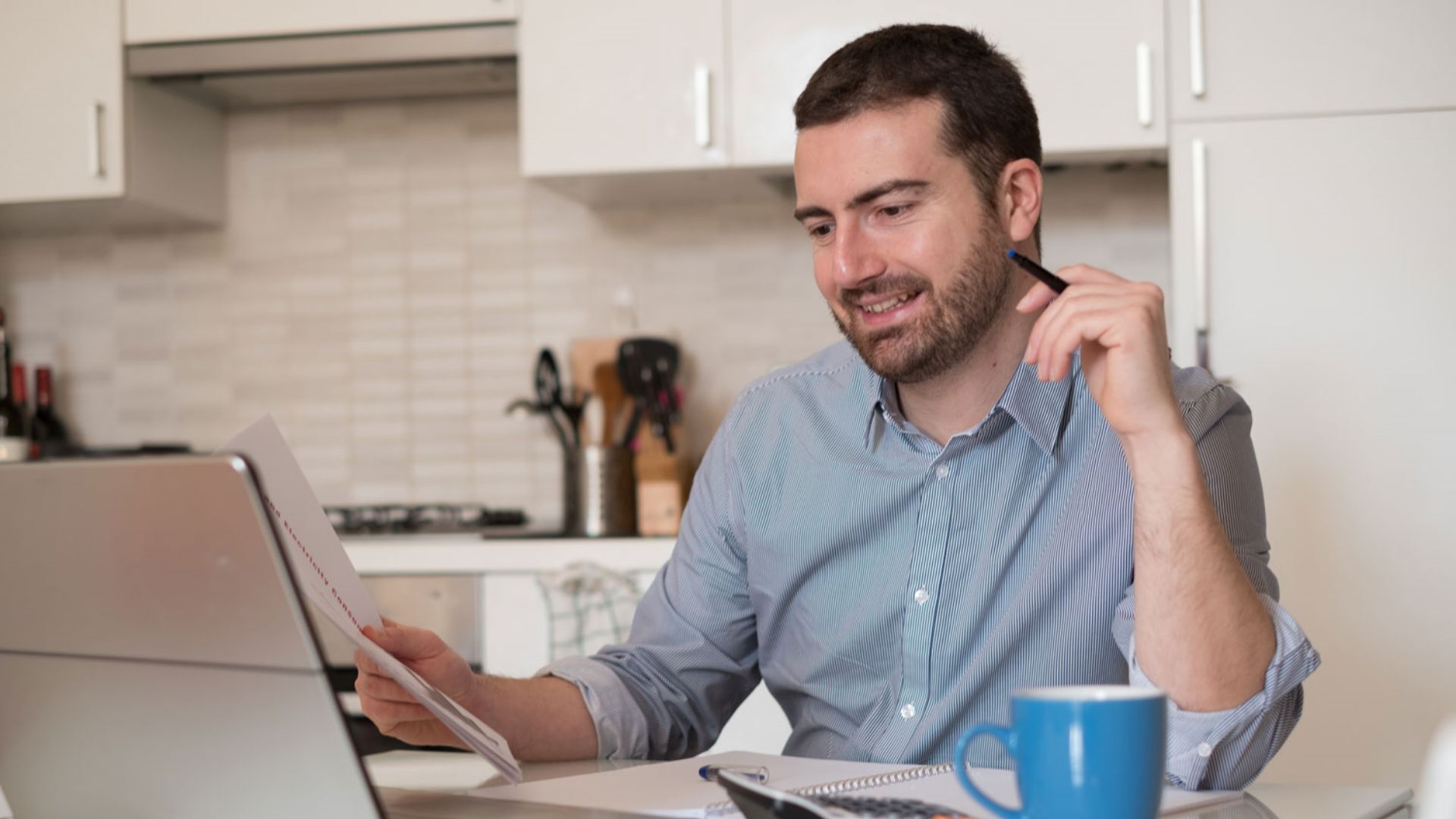 Ein Mann sitzt vor einem Laptop, daneben liegt ein Taschenrechner und in seiner Hand hält er Dokumente.