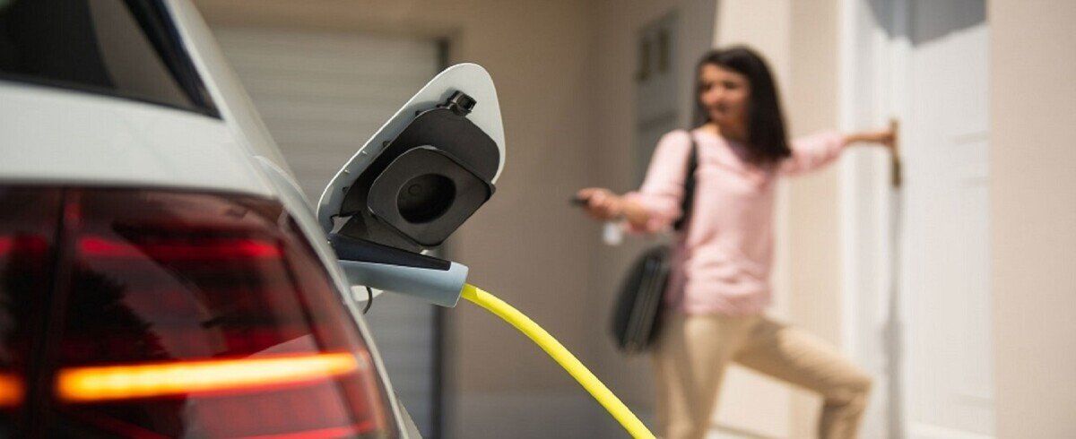 Elektroauto mit Ladekabel mit einer Frau im Hintergrund, die auf die Fernbedienung des Fahrzeugs drückt und zur Haustür geht.