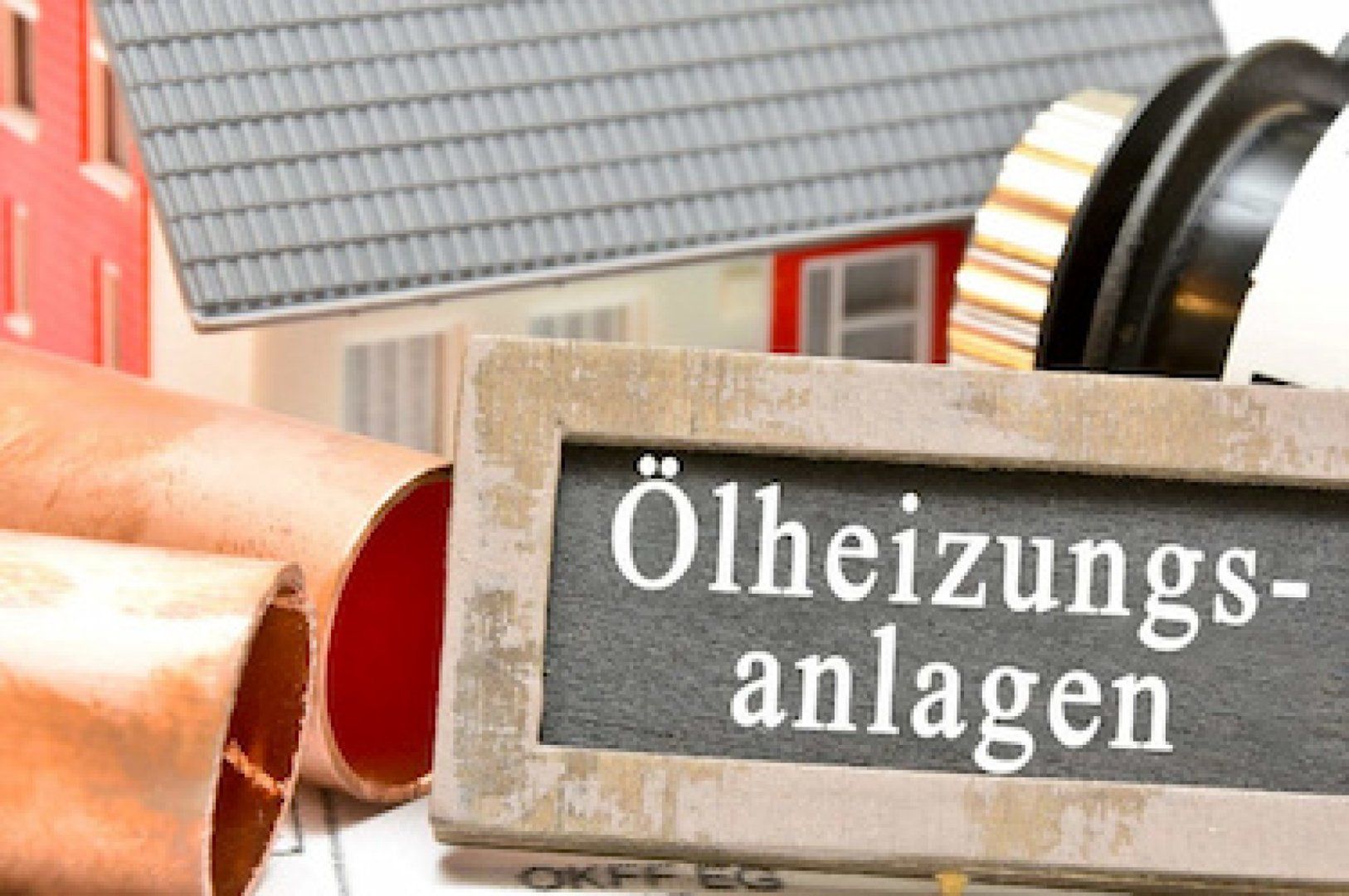 Hier ist ein kleines Haus zu sehen, davor liegen Heizungsrohre, ein Thermostatkopf und eine Tafel mit der Aufschrift „Ölheizungsanlage“.