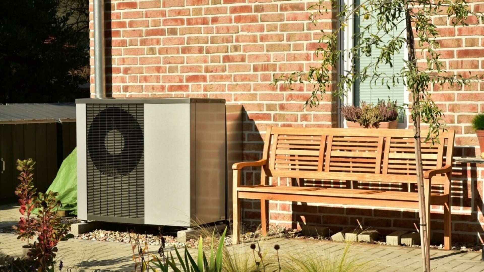 Eine Luft-Wasser-Wärmepumpe an einer Backstein-Hauswand neben einer Holzbank.