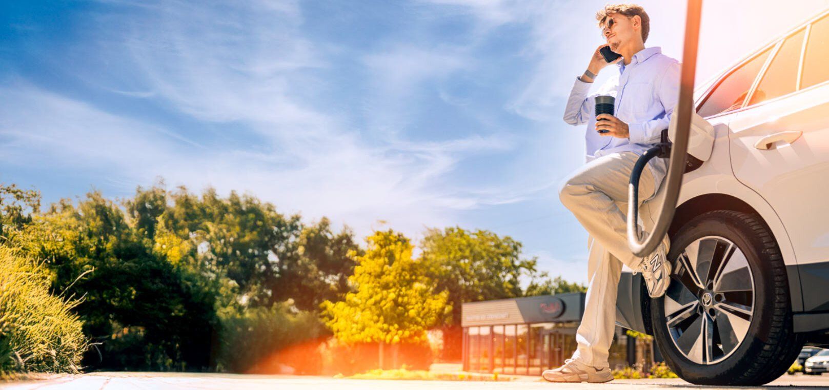 Junger Mann lehnt an E-Auto, das an Ladesäule hängt, und telefoniert