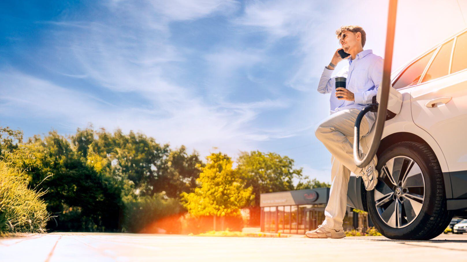 Junger Mann lehnt an E-Auto, das an Ladesäule hängt, und telefoniert