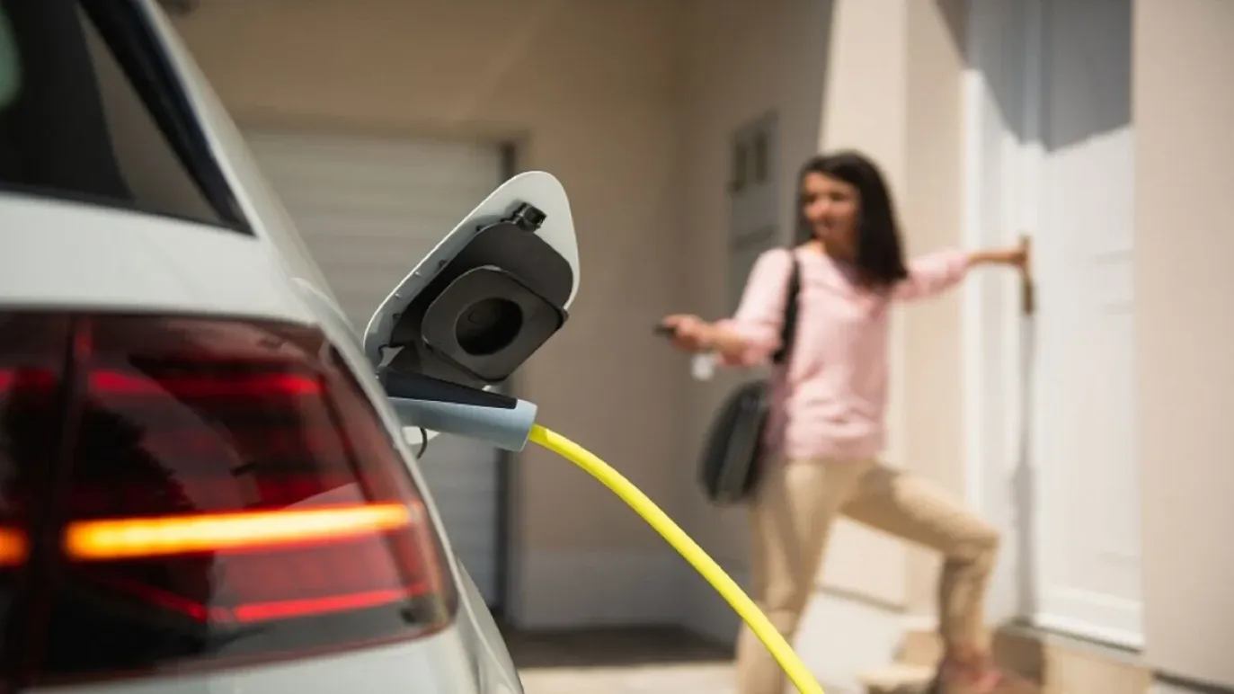 Elektroauto mit Ladekabel mit einer Frau im Hintergrund, die auf die Fernbedienung des Fahrzeugs drückt und zur Haustür geht.