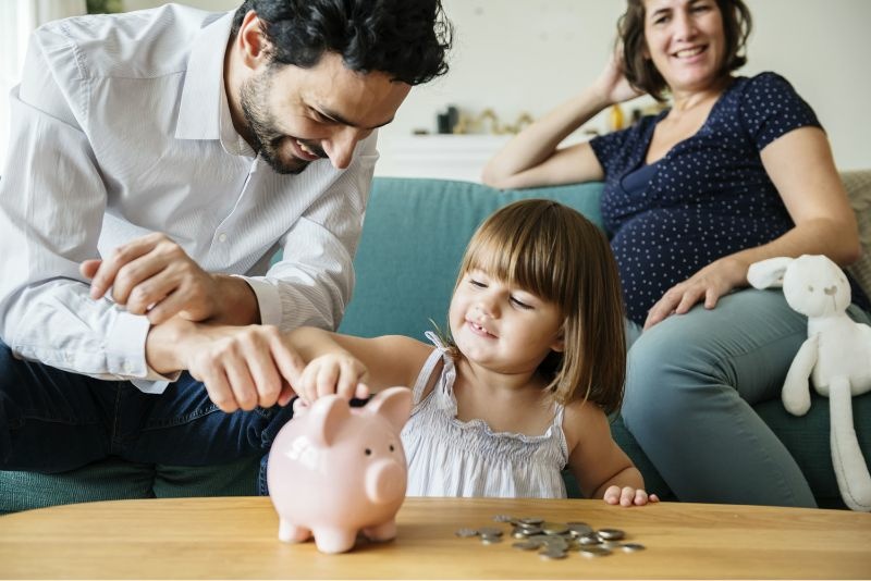 Auf dem Bild ist eine Familie mit einem Kind zu sehen. Der Vater hilft der Tochter Münzen in ein Sparschwein zu werfen. 